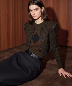 Photograph of a fair skinned woman with straight brown hair wearing a textured green jacket and black skirt on a brown carpet
