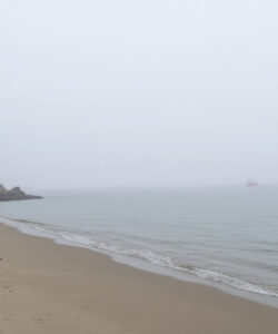 Photograph-of-a-foggy-desolate-beach-with-a-distant-lighthouse-and-a-small-boat-on-a-calm-grey-sea