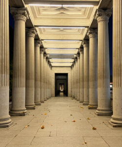 Photograph-of-a-grand-illuminated-stone-columned-promenade-at-night-with-leafy-ground-urban-backdrop-and-distant-city-lights