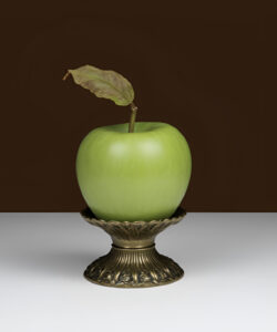 Photograph-of-a-green-apple-with-a-single-leaf-resting-on-an-ornate-bronze-stand-against-a-dark-brown-background
