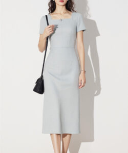 Photograph of a light skinned woman in a light blue form fitting dress holding a black shoulder bag standing against a white wall