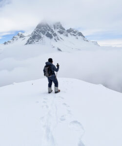 Photograph-of-a-lone-hiker-in-winter-gear-ascending-snowy-mountain-peak-with-misty-clouds-and-rugged-mountain-backdrop