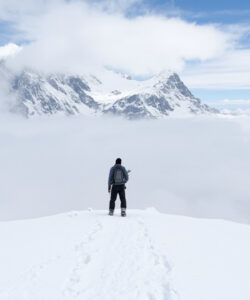 Photograph-of-a-lone-hiker-in-winter-gear-standing-on-a-snowy-ridge-with-majestic-mountains-and-clouds-in-background