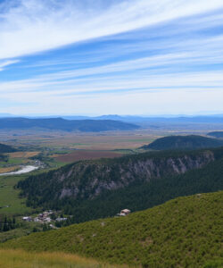 Photograph-of-a-lush-green-valley-with-rolling-hills-winding-road-scattered-houses-and-distant-mountains-under-a-blue-sky