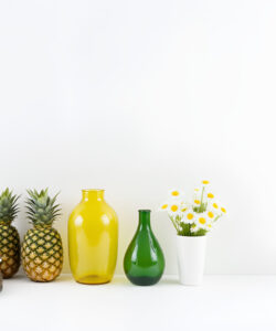 Photograph-of-a-minimalist-white-background-with-a-yellow-vase-green-vase-pineapples-grey-shoes-yellow-flowers-and-a-green-pot