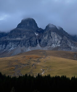 Photograph-of-a-misty-jagged-mountain-peak-under-cloudy-sky-with-a-winding-path-through-autumnal-forested-hills-below