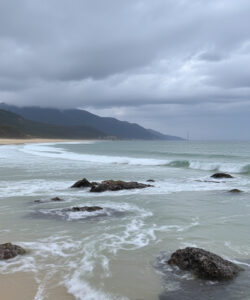 Photograph-of-a-misty-overcast-beach-with-rocky-shores-gentle-waves-and-distant-forested-mountains-under-a-cloudy-sky