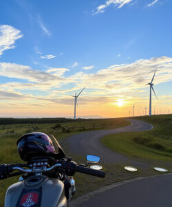 Photograph-of-a-motorcycle-s-handlebar-at-sunset-with-wind-turbines-in-the-background-winding-road-and-clear-blue-sky