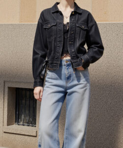 Photograph of a pale skinned brunette woman in a black denim jacket black lace bra and high waisted blue jeans