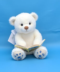 Photograph-of-a-plush-white-teddy-bear-with-blue-paw-prints-sitting-and-reading-a-book-against-a-blue-backdrop