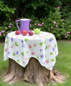 Photograph-of-a-purple-watering-can-red-apples-and-green-bowl-on-a-floral-tablecloth-over-a-tree-stump