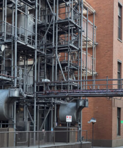 Photograph-of-a-red-brick-building-with-complex-industrial-metal-scaffolding-and-large-pipes-urban-setting-with-a-narrow-street