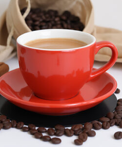Photograph-of-a-red-ceramic-cup-of-frothy-latte-on-a-saucer-surrounded-by-scattered-coffee-beans-star-anise-and-cloth