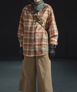 Photograph of a red haired person in plaid shirt tan pants black gloves and hat standing against a dark background