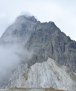Photograph-of-a-rugged-mist-covered-mountain-peak-with-rocky-textures-partially-obscured-by-clouds-tiny-hikers-visible-near-base