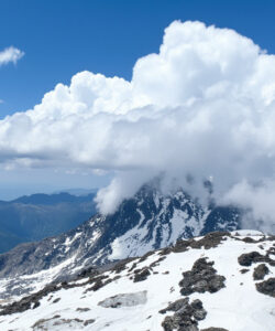 Photograph-of-a-rugged-snow-covered-mountain-peak-shrouded-in-clouds-with-distant-blue-green-valleys-and-clear-blue-sky