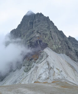Photograph-of-a-rugged-snow-covered-mountain-peak-with-a-cloud-partially-obscuring-the-summit-under-a-partly-cloudy-sky