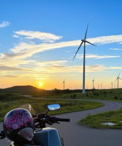 Photograph-of-a-scenic-coastal-road-with-wind-turbines-at-sunset-featuring-a-helmeted-motorcyclist-and-lush-green-landscape