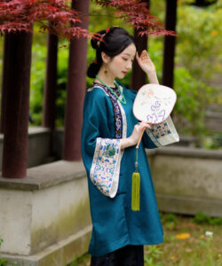 Photograph of a serene asian woman in traditional teal robes holding a fan with delicate floral embroidery under a tree