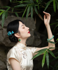 Photograph of a serene asian woman with black hair in a bun wearing a cream cheongsam touching bamboo leaves