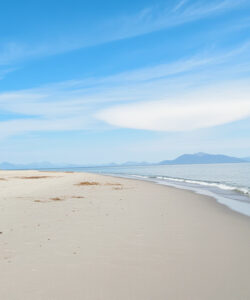 Photograph-of-a-serene-beach-with-a-tall-lighthouse-on-the-left-under-a-clear-blue-sky-with-wispy-clouds