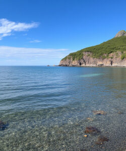 Photograph-of-a-serene-beach-with-clear-turquoise-water-rocky-shore-and-a-lush-green-mountainous-backdrop-under-a-blue-sky