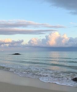 Photograph-of-a-serene-beach-with-gentle-waves-clear-blue-sky-fluffy-white-clouds-and-a-few-distant-rocks