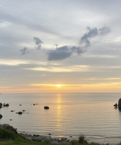 Photograph-of-a-serene-coastal-landscape-at-sunset-featuring-calm-waters-rocky-cliffs-and-lush-greenery-with-a-cloudy-sky