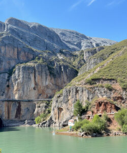 Photograph-of-a-serene-mountainous-landscape-with-a-turquoise-river-rocky-cliffs-a-green-roofed-building-and-a-waterfall