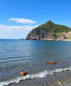 Photograph-of-a-serene-pebble-beach-with-clear-turquoise-water-green-hills-and-a-few-clouds-in-a-bright-blue-sky