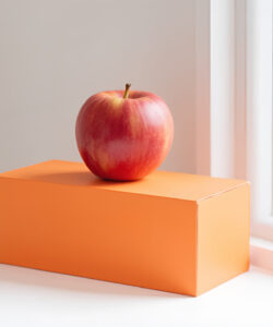 Photograph-of-a-single-red-apple-atop-an-orange-rectangular-box-on-a-white-windowsill-with-a-bright-window