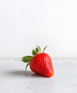 Photograph-of-a-single-vibrant-red-strawberry-with-green-leaves-placed-on-a-clean-white-wooden-surface-against-a-plain-background