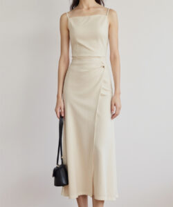 Photograph of a slender asian woman in a cream sleeveless midi dress holding a black crossbody bag light wooden floor