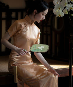 Photograph of a slender asian woman in a gold silk cheongsam holding a green fan against a dimly lit background