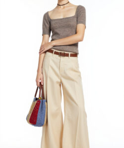 Photograph of a slender androgynous person with short dark hair wearing a grey top beige high waisted pants and a blue bag