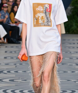 Photograph of a slender blonde model with pale skin walking on a runway in a white oversized t shirt and fringe skirt