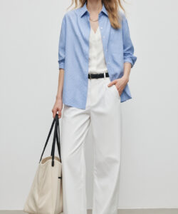Photograph of a slender blonde woman in a blue button up shirt white pants black belt beige handbag and sandals