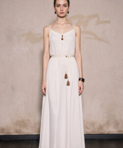 Photograph of a slender fair skinned woman in a white floor length dress with thin straps against a textured beige wall