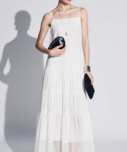 Photograph of a slender fair skinned woman in a white sheer maxi dress holding a black clutch against a plain background