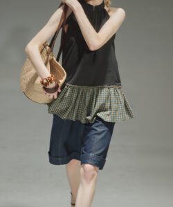 Photograph of a slender light skinned woman in a brown cap sleeveless black top plaid skirt denim shorts and loafers