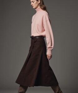Photograph of a slender light skinned woman in a pale pink sweater and brown high waisted skirt walking against a gray background