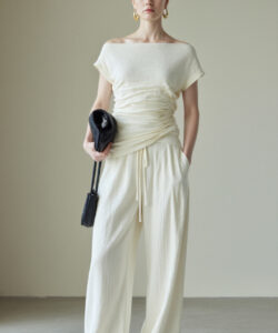 Photograph of a slender light skinned woman in an off shoulder cream colored ruched top and loose pants holding a black clutch