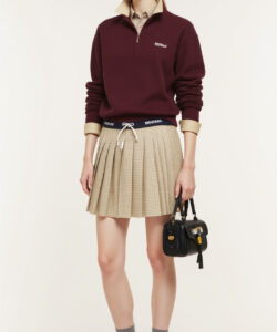 Photograph of a slender light skinned woman with glasses wearing a burgundy sweater beige pleated skirt grey socks and black shoes