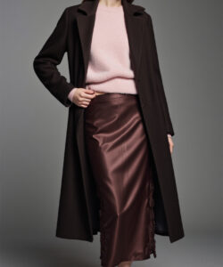 Photograph of a slender light skinned woman with long brown hair wearing a pink sweater brown coat and brown satin skirt