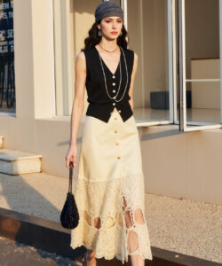 Photograph of a slender light skinned woman with long dark hair wearing a black vest and beige lace skirt carrying a purse