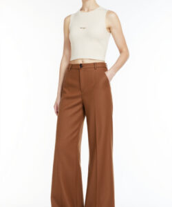 Photograph of a slender light skinned woman with short brown hair wearing a cream sleeveless crop top and high waisted brown pants