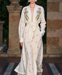 Photograph of a slender woman in a cream robe with floral embroidery walking on a stone pathway