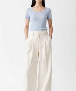 Photograph of a slender woman in a light blue t shirt beige pants and a straw hat standing against a plain background
