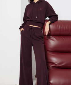 Photograph of a slender woman in a maroon hoodie and matching loose pants leaning against a red leather couch