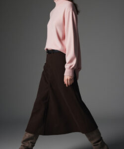 Photograph of a slender woman in a pink turtleneck brown high waisted skirt and grey leg warmers walking against a grey background
